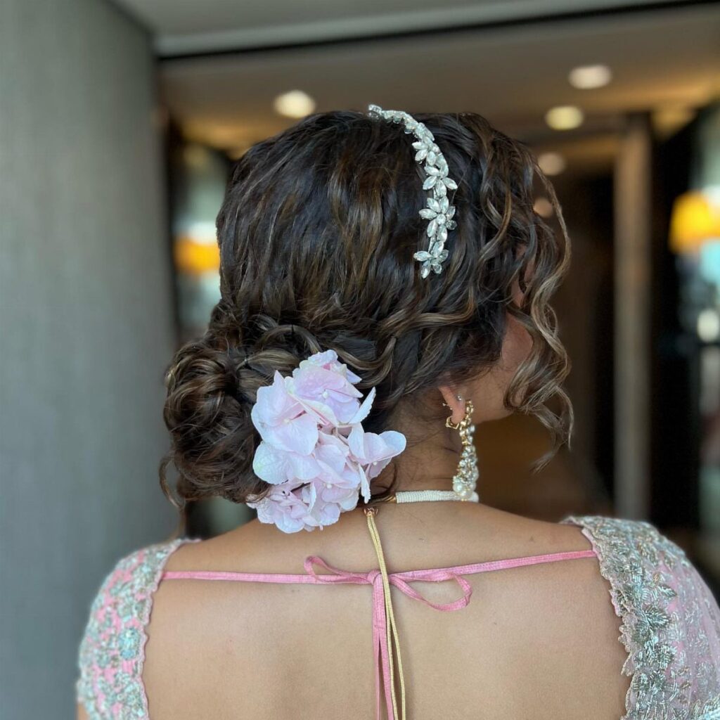 Low curly hair bun decorated with a pink flower and silver headband.