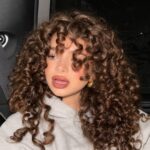 Woman with voluminous brown curly hair and bangs wearing a grey hoodie.