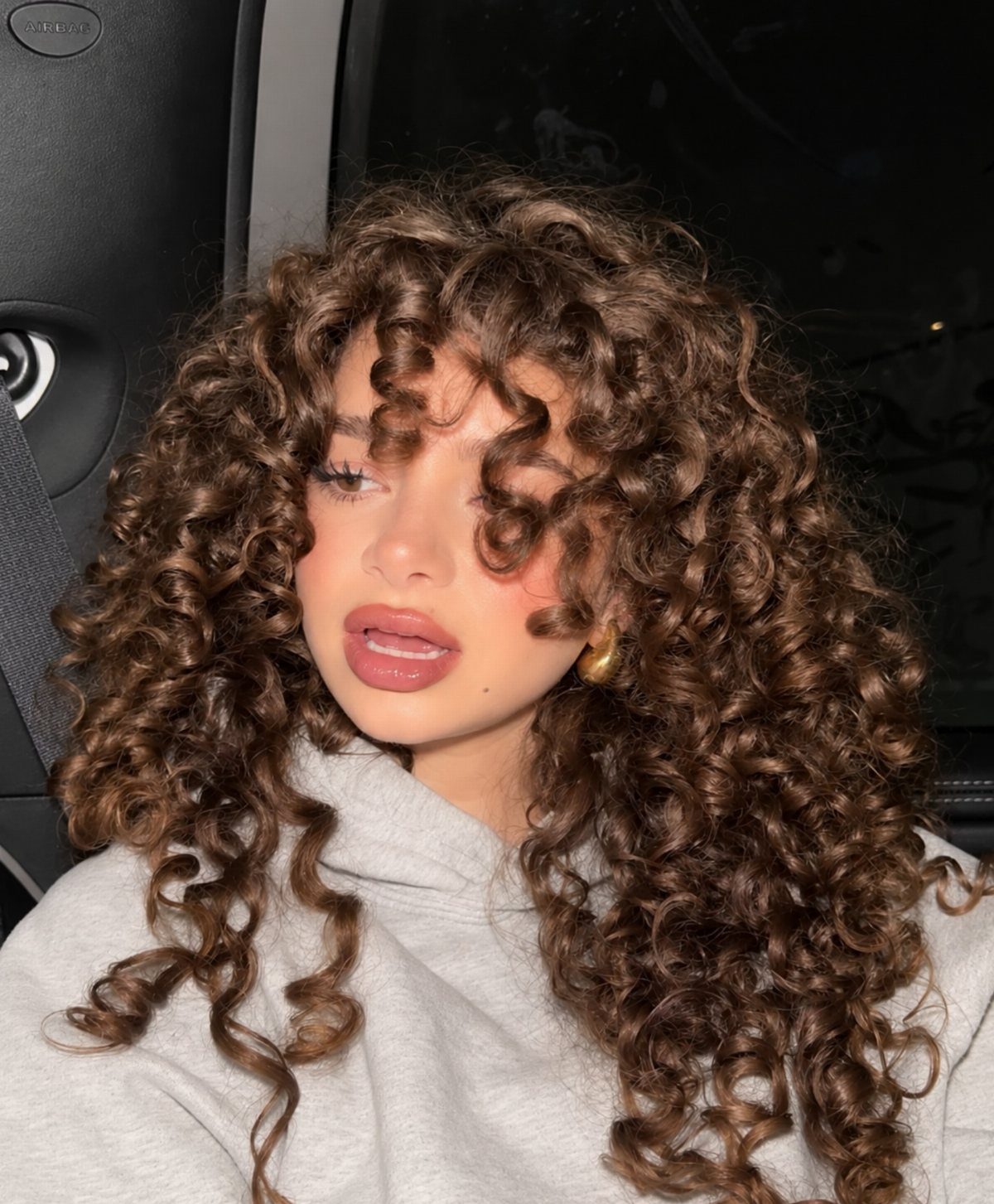 Woman with voluminous brown curly hair and bangs wearing a grey hoodie.