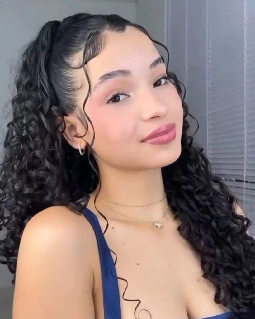 Young woman with long dark curly hair styled in a high ponytail.