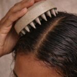Hand using a beige spiked scalp massager on dark hair at the part.