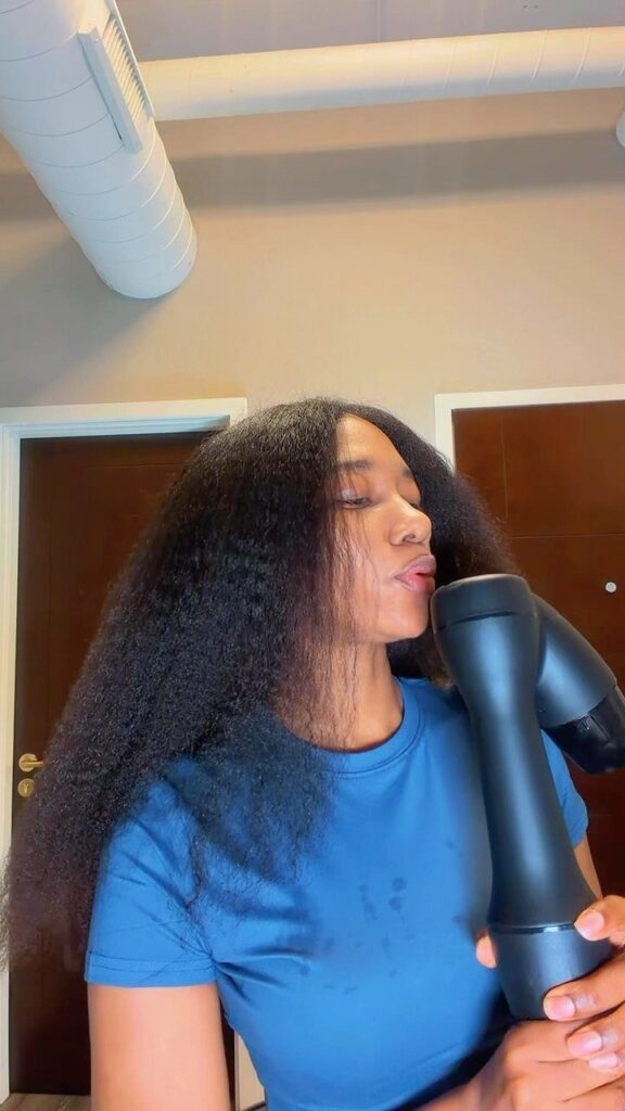 Woman with voluminous natural hair using a black hair dryer in a room.