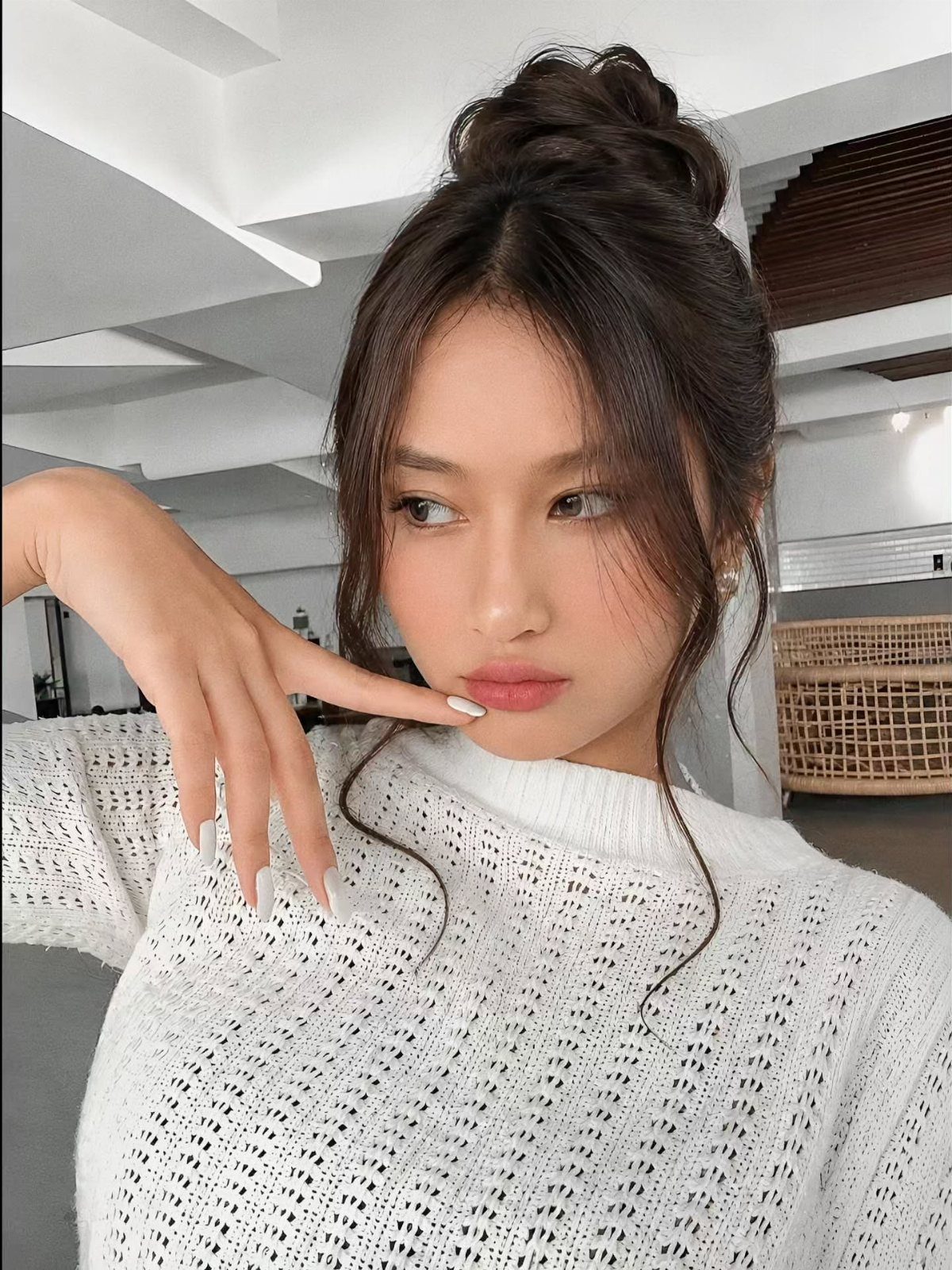 Woman with a high bun hairstyle wearing a white knitted sweater posing indoors.
