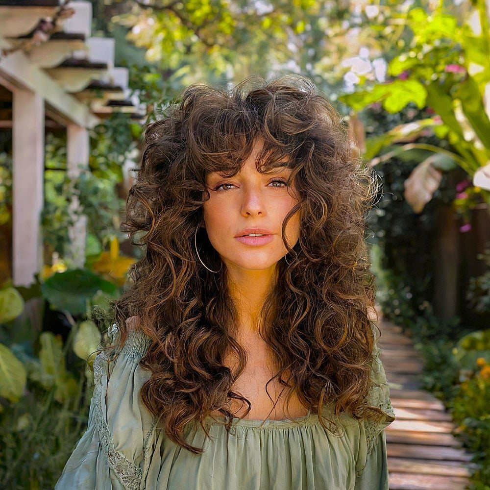 Woman with long brown curly hair and curly bangs in a lush garden